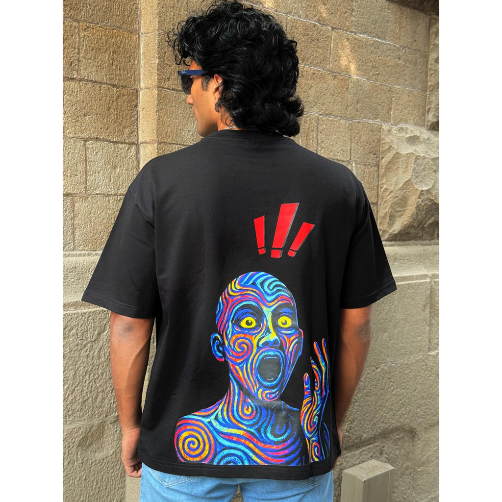 Person wearing a black t-shirt with a colorful graphic design on the back, standing against a stone wall.